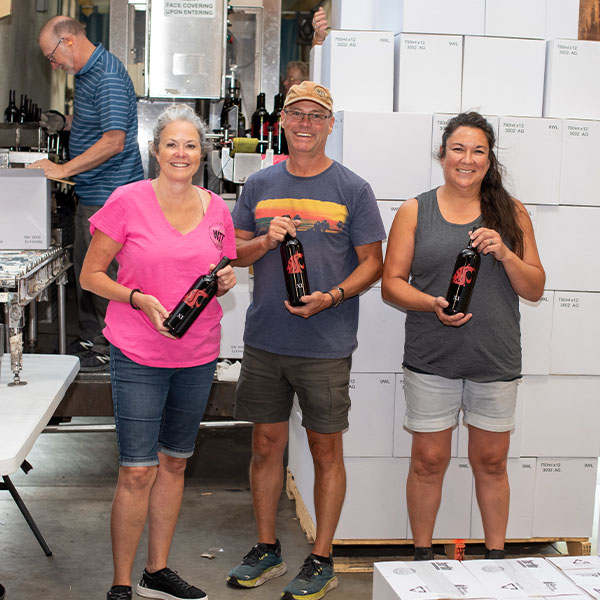 Three people holding wine bottles in warehouse.