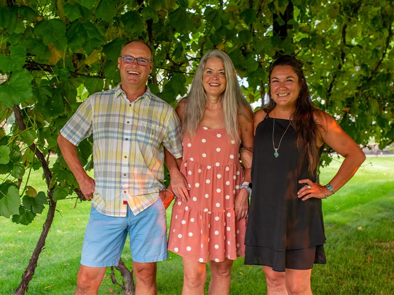 Three people smiling outdoors under greenery