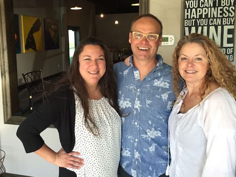 Three people smiling indoors near wine sign.