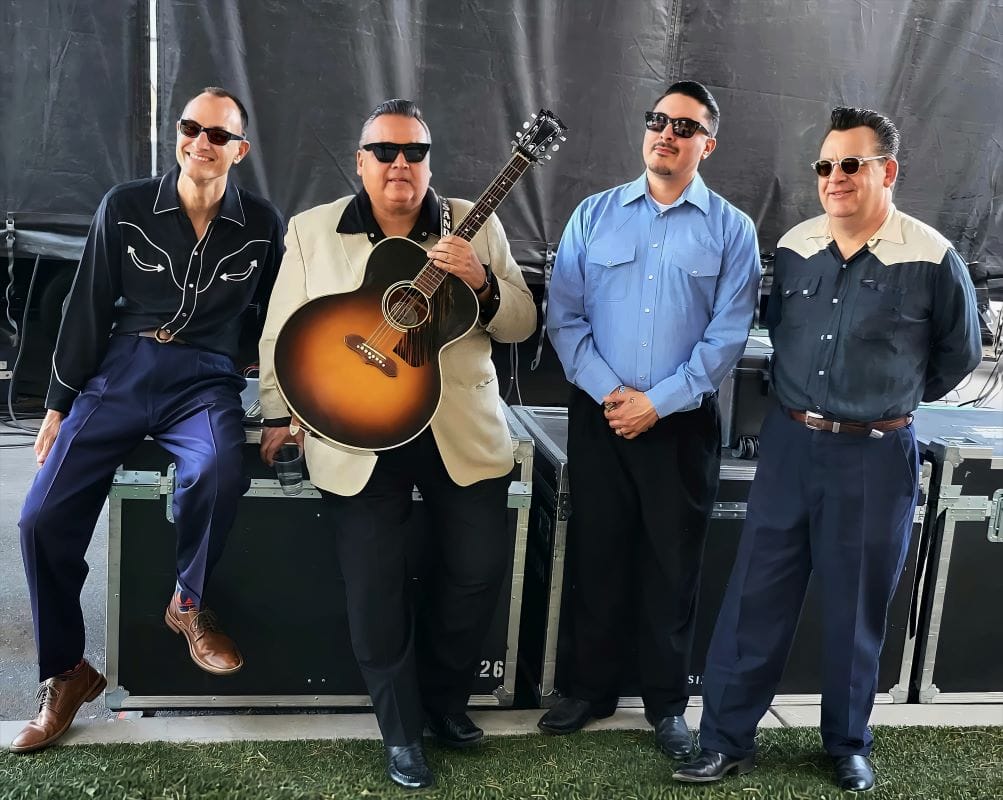 Four musicians posing with a guitar backstage.