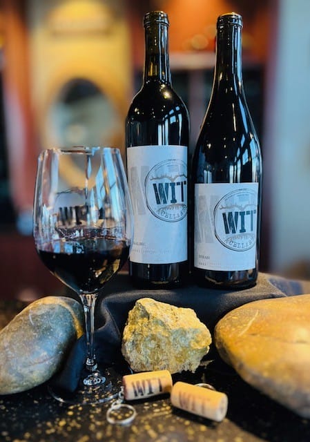 May-2025-Release-Weekend Red wine with bottles and glass on rocky table.