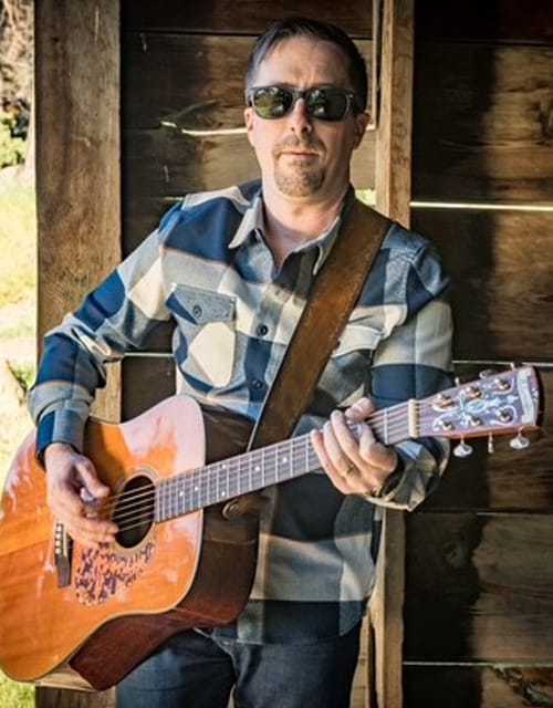 Man playing guitar outdoors in sunglasses.