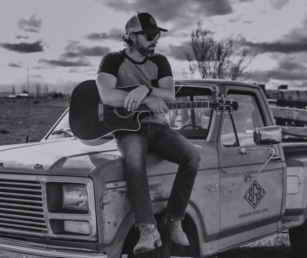 Zac-Grooms Man sitting on truck with guitar, rural setting.