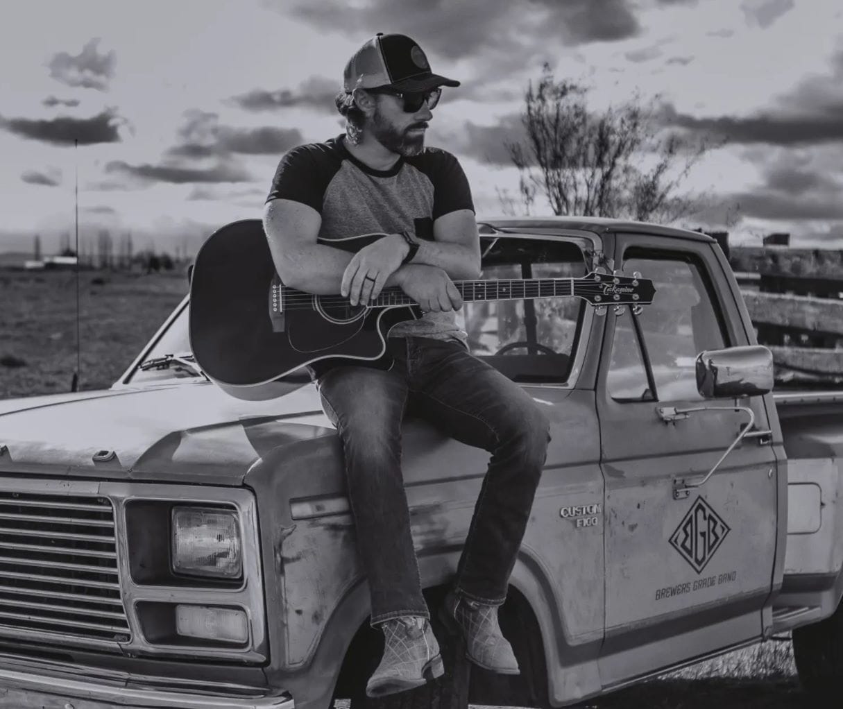 Man sitting on truck with guitar, rural setting.