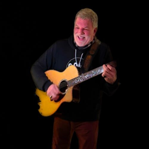 Man playing guitar, smiling on dark background.