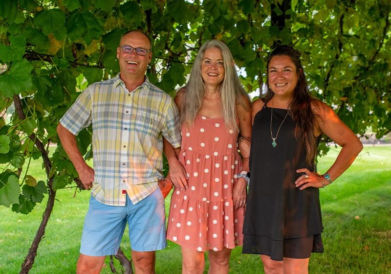 Three people smiling outdoors under greenery
