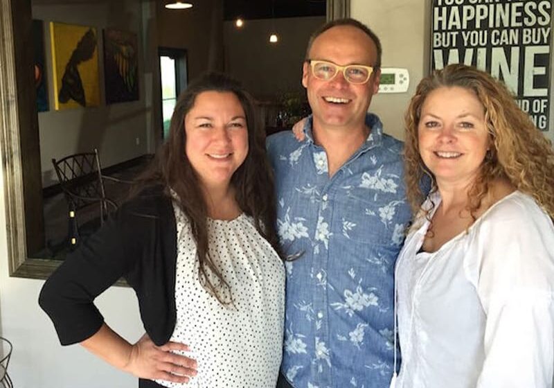 Three people smiling indoors near wine sign.
